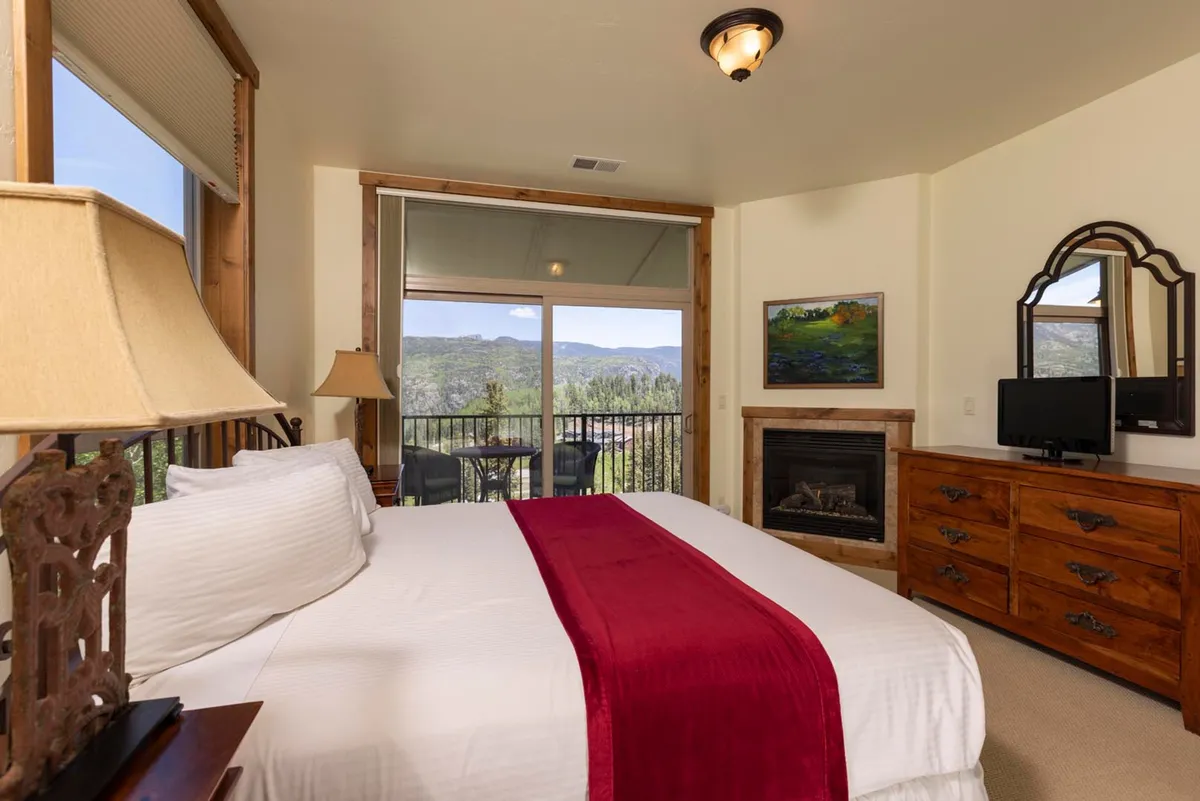 A restful bedroom with a comfortable bed adorned with a red accent, expansive windows showcasing mountain vistas, a fireplace, and tasteful wooden furniture. The space radiates calm and provides a perfect setting for restful nights or reading by the fire.