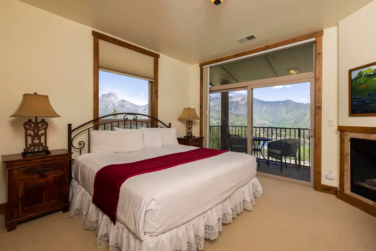 A cozy bedroom featuring a plush bed with a vibrant red throw, a nightstand with a lamp, and a fireplace. Large windows reveal breathtaking mountain views, creating a connection to the alpine surroundings. The decor is inviting, and the setting suggests deep relaxation after a day outdoors.