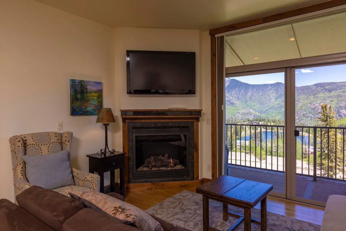 A warm, welcoming living room centered around a glowing fireplace, with comfortable seating and large windows that frame stunning mountain and lake views. The space invites small groups or families to gather, relax, and share stories after a day of resort activities.