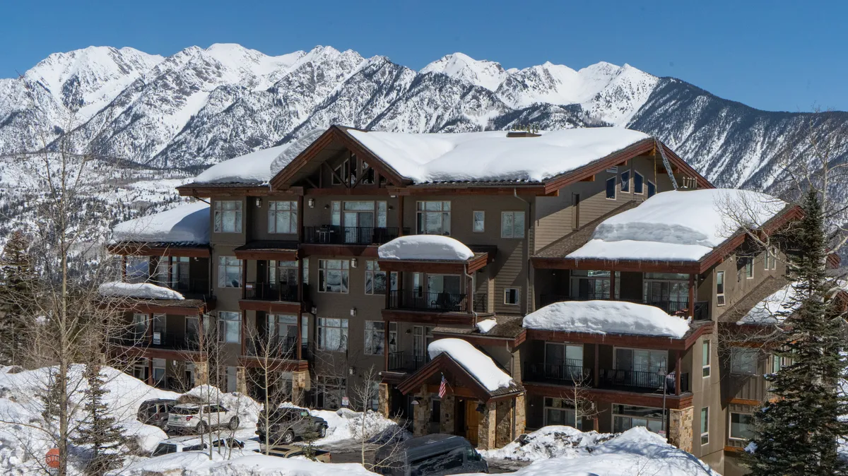 An exterior perspective showcases the building’s snow-draped balconies and distinctive architecture, with towering mountains as a majestic backdrop. The setting feels both cozy and adventurous, highlighting the property’s unbeatable location.