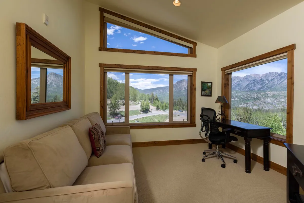 A tranquil living area with a modern desk and comfortable sofa positioned beside expansive windows that frame sweeping mountain views. Natural light floods the space, inviting relaxation or quiet productivity while the beauty of the outdoors remains ever-present.