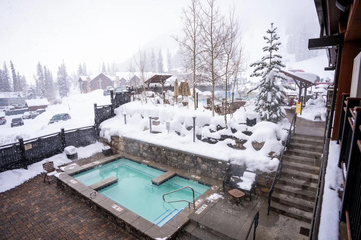 A snow-covered resort landscape with an inviting outdoor hot tub surrounded by pine trees. Guests are visible relaxing in the steaming water, while others play in the snow nearby. The stone wall and steps add to the sense of a shared, welcoming outdoor space perfect for winter fun.