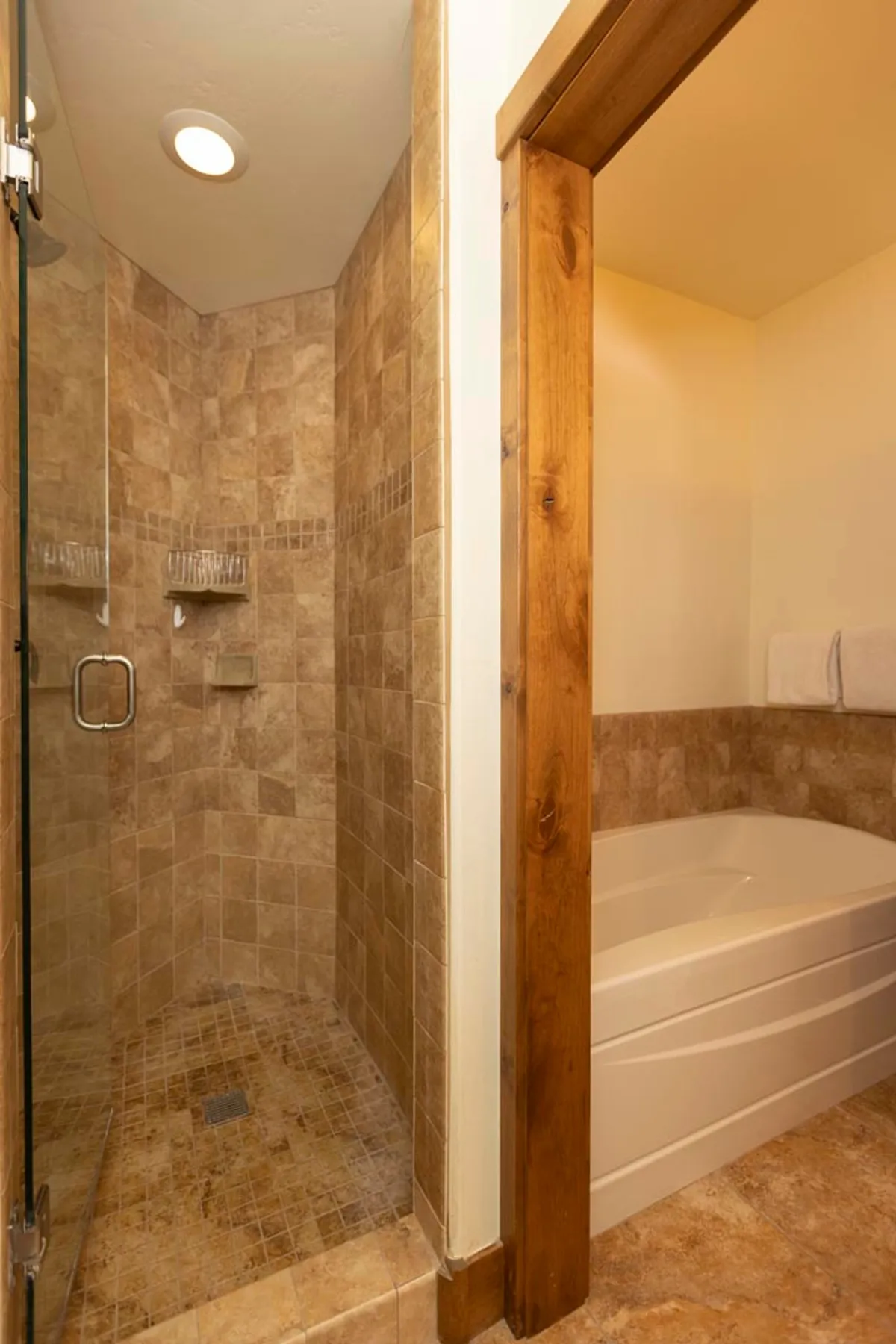 A modern bathroom with a glass-enclosed shower, inviting bathtub, textured tiles, and a wooden door frame. The tranquil atmosphere is enhanced by thoughtful design details, offering a serene space for relaxation and self-care.