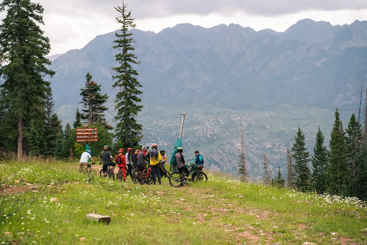 A lively group of cyclists takes a break on a mountain trail, surrounded by a dramatic mountain range. The signpost hints at multiple trail options, and the expansive alpine landscape provides a sense of adventure and camaraderie. The scene is full of fresh mountain air and excitement, perfectly capturing the spirit of summer mountain biking and hiking.