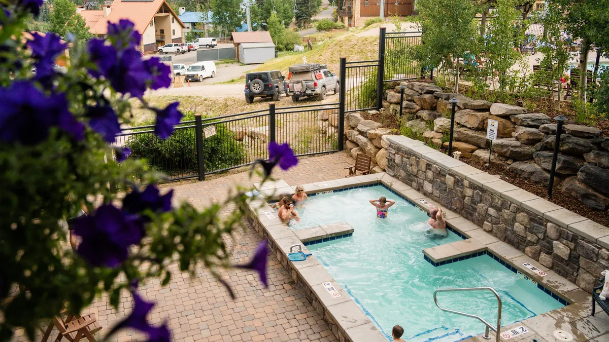 A vibrant outdoor swimming pool is framed by blooming summer flowers and a stone wall. People are seen enjoying the water and lounging in the sun, creating a joyful and relaxed atmosphere. The scene evokes the carefree enjoyment of summer days in a mountain setting.