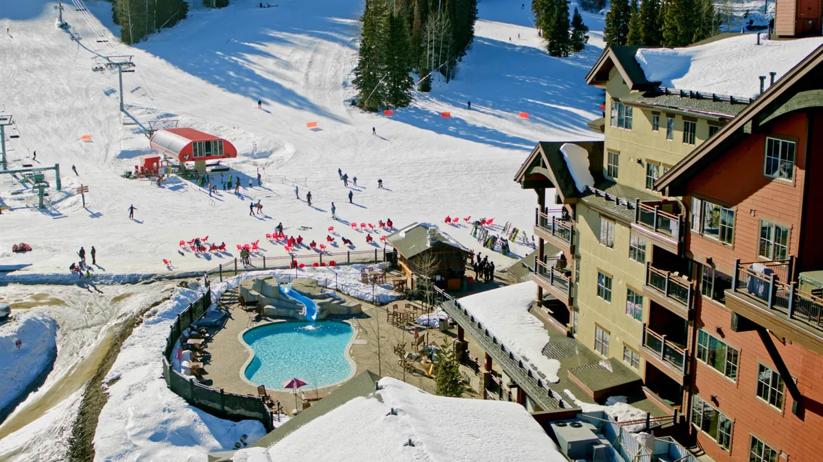 The image offers a wide view of ski slopes and a nearby pool area, with a ski lift visible in the background. Snow blankets the ground and trees, while people can be seen enjoying various activities, from skiing to lounging by the pool.