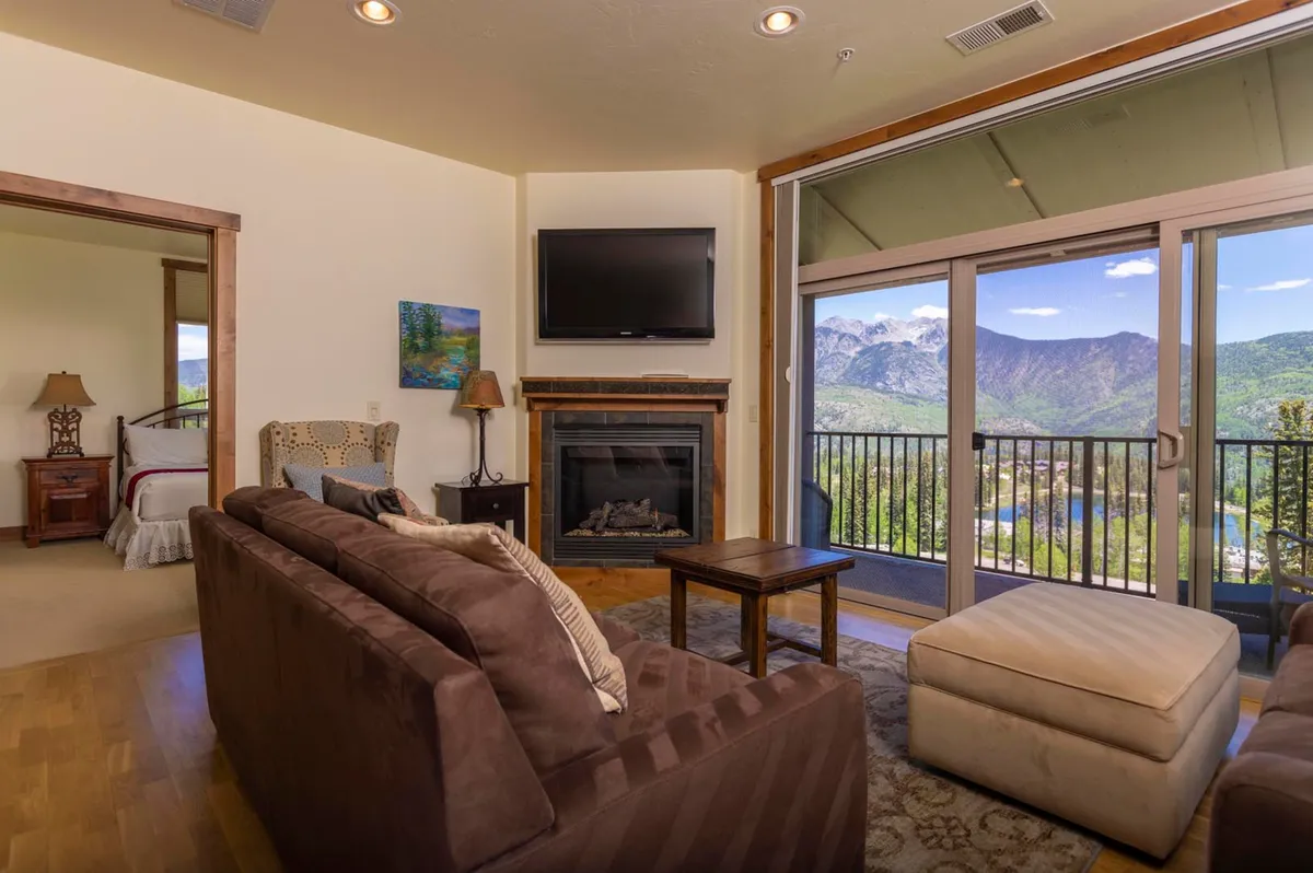 A spacious, softly-lit living room with plush sofas and armchairs arranged around a modern fireplace. Expansive windows dominate one wall, revealing breathtaking views of the Needles Mountains. The seating is inviting, perfect for gatherings, and there are touches of tasteful artwork on the walls, with a warm, welcoming atmosphere throughout.
